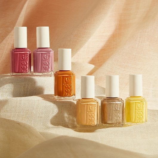 Six bottles with shades of yellow, orange and pink nail polish against a beige linen background