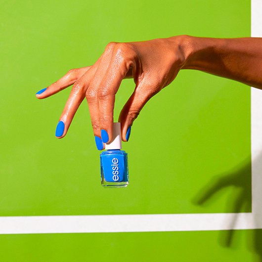 Hand with blue nails holding a bottle of nail polish in the same color on a bright green background