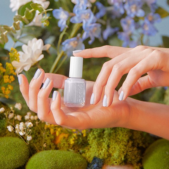Two hands with nails painted blue holding a bottle of nail polish in the same shade with flowers and moss in the background