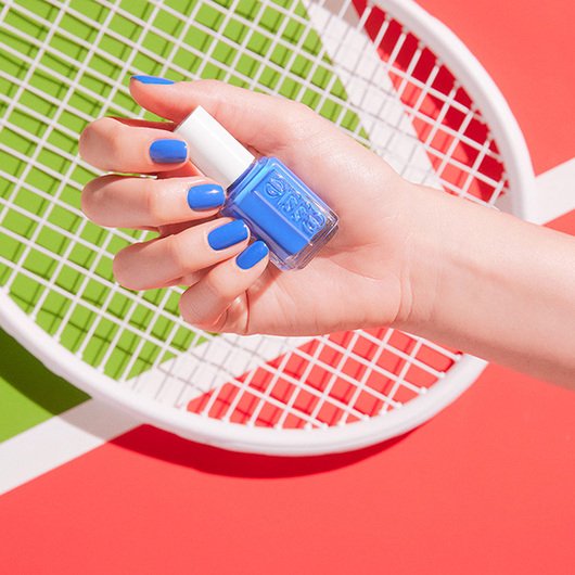 Hand holding a bottle of nail polish in the shade push play with nails painted in the same color with a tennis court and racket background