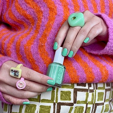 Light skin model wearing very colorful clothing and statement rings holding green essie nail polish