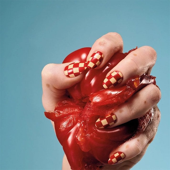 Light skin hand wearing red and beige checkered nail art squeezing a large juicy red tomato