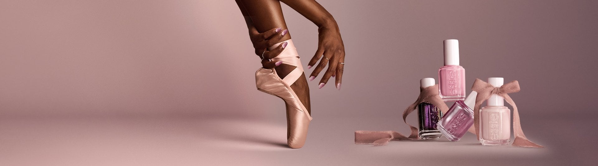 Three essie ""Ballet Slippers"" nail polish bottles tucked inside matching satin pointe ballet shoes against a pink background.