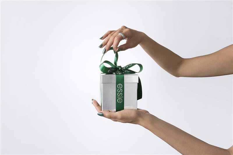 A silver gift box tied with a green "essie" branded ribbon being opened by a hand with dark green nails.