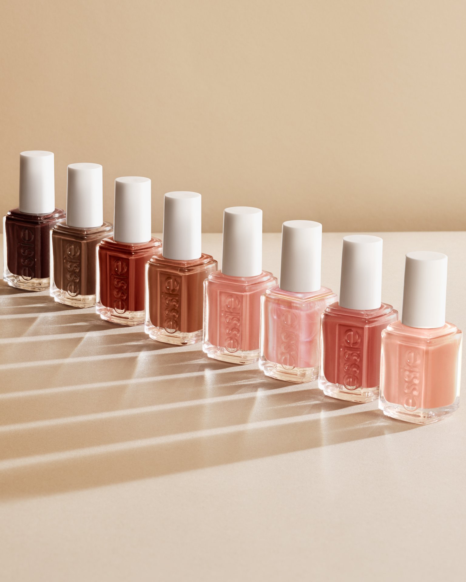 A line of eight essie nail polish bottles ranging from deep espresso to pale peach, casting long shadows on a beige surface.