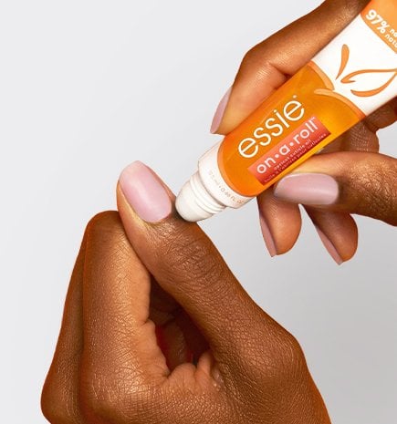 A hand applying essie "On A Roll" apricot cuticle oil from a roll-on tube to the base of a fingernail.