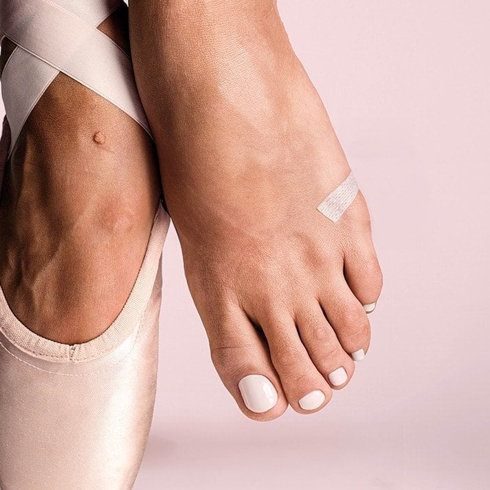 Close-up of essie Ballet Slippers nail polish on toes, paired with a satin ballet slipper and pink ribbons to highlight the classic sheer pink color.