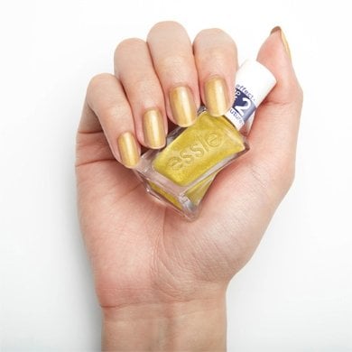 A hand holds a bottle of metallic gold polish labeled "Essie." The nails are painted in matching shade
