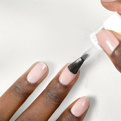 The tips of three dark skin fingers wearing light pink polish getting a top coat brushed on a nail