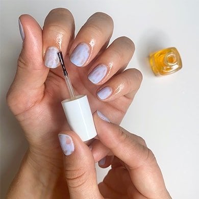 Bottle of cuticle oil next to light skin hands, one brushing the oil onto cuticles.