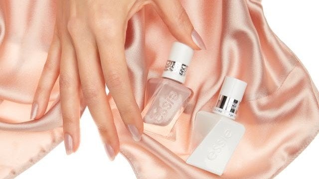 a hand with light pink painted nails holding a soft pink essie gel couture polish bottle laying next to a gel top coat bottle