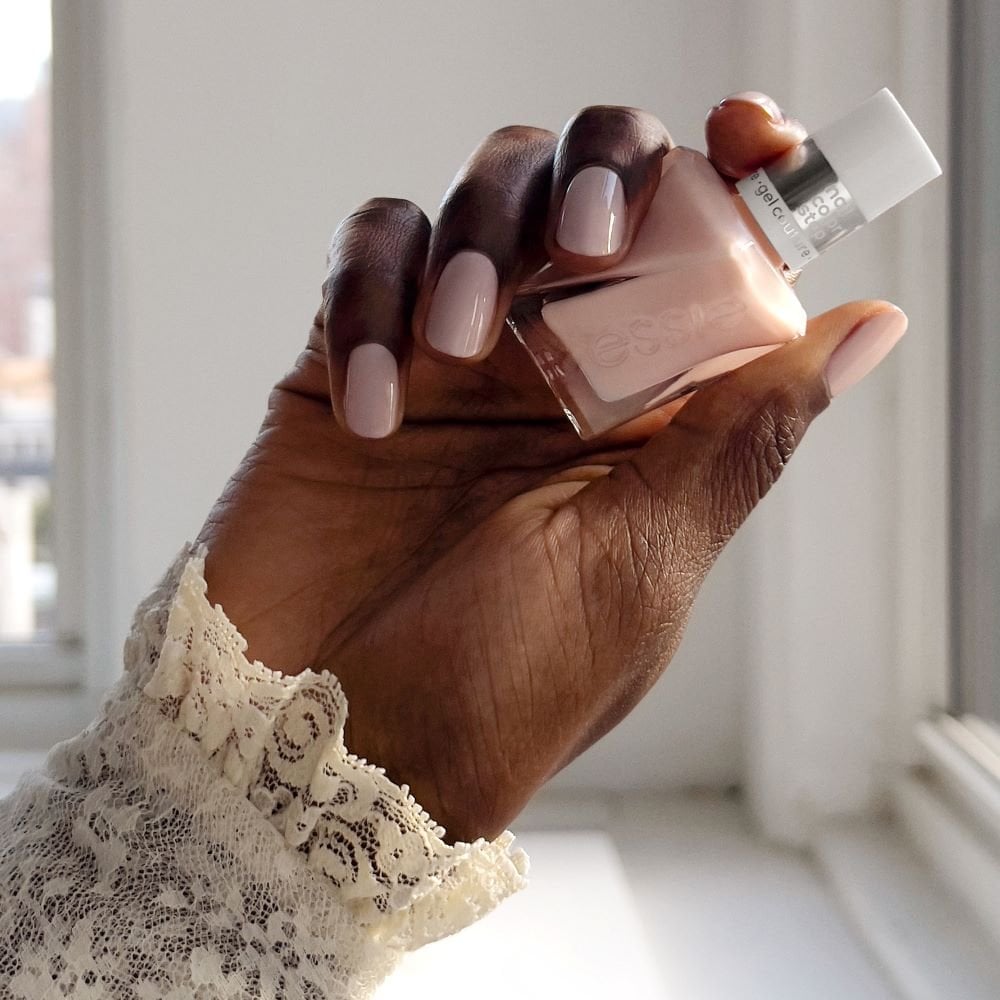 a hand with light pink painted nails and a lace sleeve hold a light pink bottle of essie gel couture polish