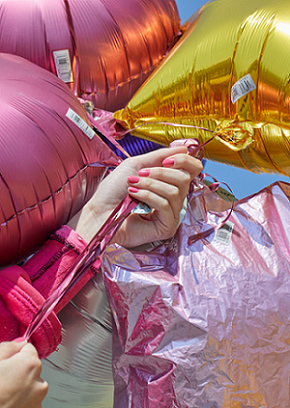 a hand with pink painted nails holding several metalllic pink & yellow balloons with blue sky background
