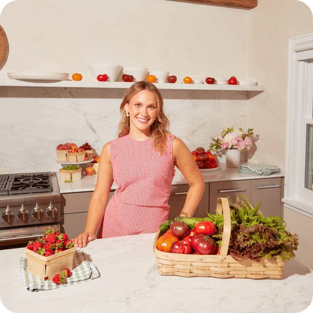 Woman in red gingham outfit smiling in a white kitchen with baskets of fresh vegetables, fruit and flowers