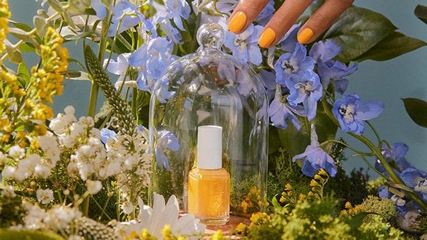 A hand reaching to lift a clear glass cloche that contains a bottle of nail polish surrounded by flowers