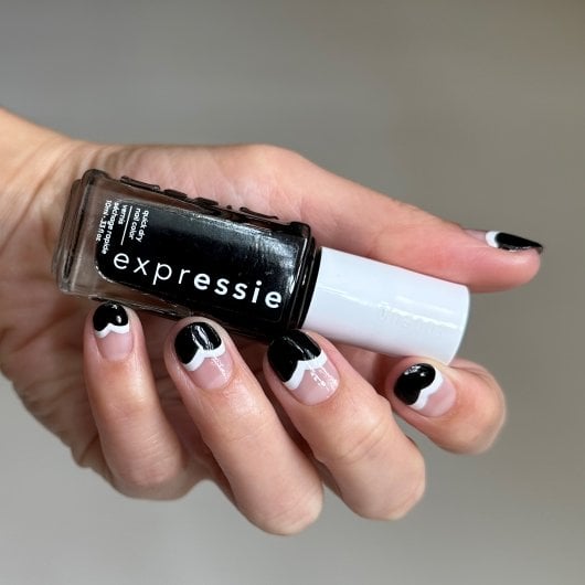 hand with black and white nail art on each fingernail holds a black bottle of nail polish 