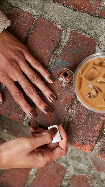 model in cream sweater painting nails with essie mocha mousse nail polish next to an iced coffee
