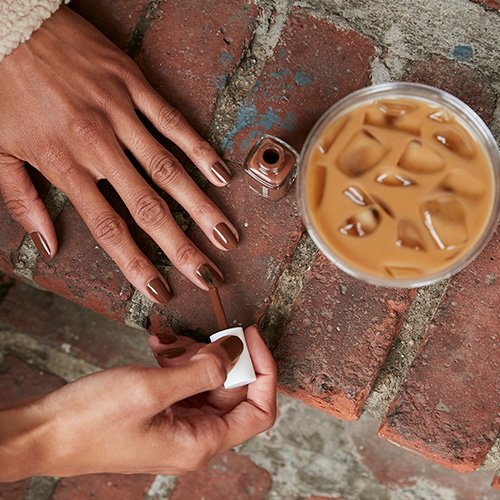 model painting nails with essie mocha mousse nail polish next to an iced coffee