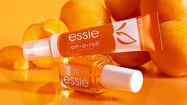 two essie apricot cuticle oils (one in a roller and one in a bottle) stacked on top of one another on an orange surface with apricots in the background