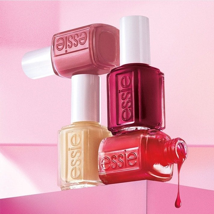 Four essie nail polish bottles stacked on pink poduim; a single pink polish drop falls from an open, sideways bottle.