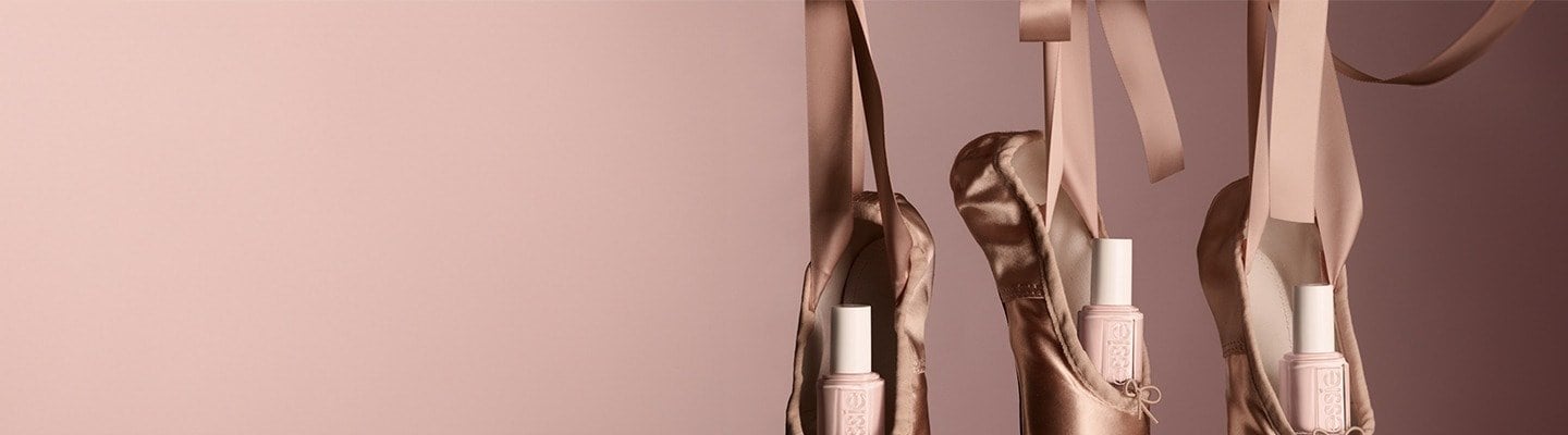 Three essie ""Ballet Slippers"" nail polish bottles tucked inside matching satin pointe ballet shoes against a pink background.