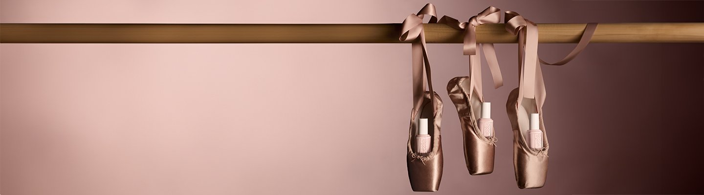 Three essie ""Ballet Slippers"" nail polish bottles tucked inside matching satin pointe ballet shoes against a pink background.