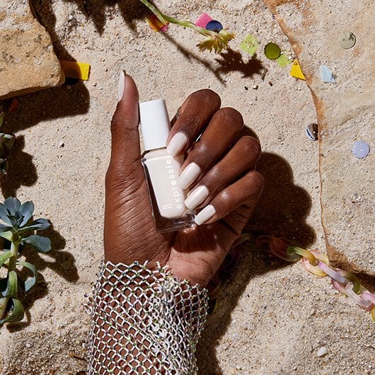 Model's white manicured hand holding white nail polish bottle in front of a rocky beach background